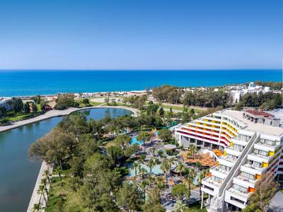 Hotelový komplex s barevnou budovou, bazénem, jezírkem a výhledem na modré moře pod jasnou oblohou.