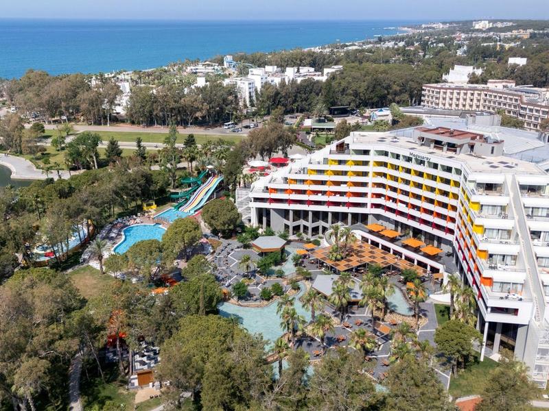 Velký hotel s zakřiveným designem, obklopený zahradami a bazénem, blízko moře.