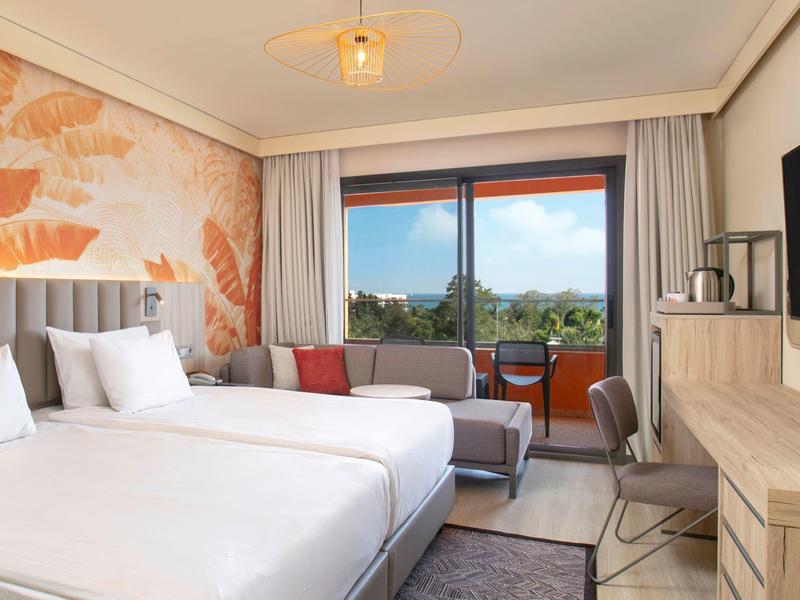 Světý hotelový pokoj se dvěma postelemi, balkonem a moderním zařízením s výhledem na přírodu.