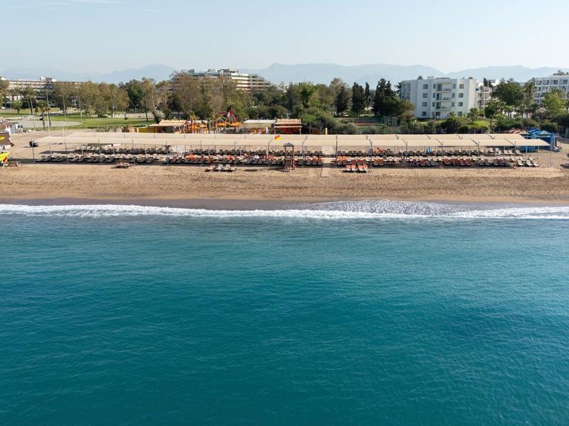 Pláž s mnoha slunečníky a lehátky, budovy a zelené stromy v pozadí.