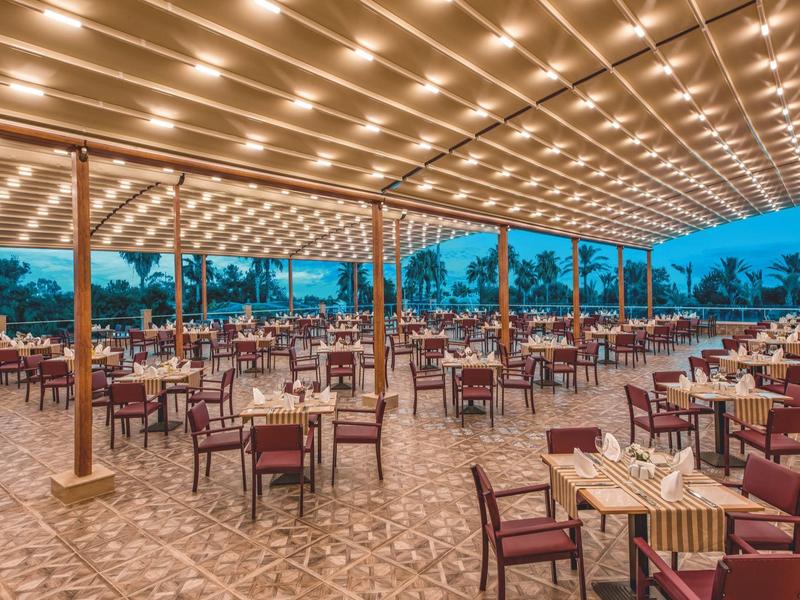 Ristorante all'aperto con molti tavoli, sedie rosse e luci a stringa sotto un tetto al crepuscolo.