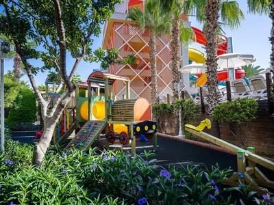 Bunter Kinderspielplatz mit Rutsche, Klettergerüst und Wippe umgeben von Palmen und Pflanzen.