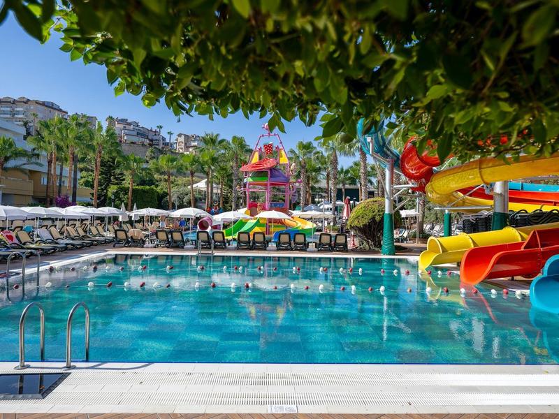 Großes Schwimmbecken mit Wasserrutschen, Sonnenliegen und Palmen unter blauem Himmel.