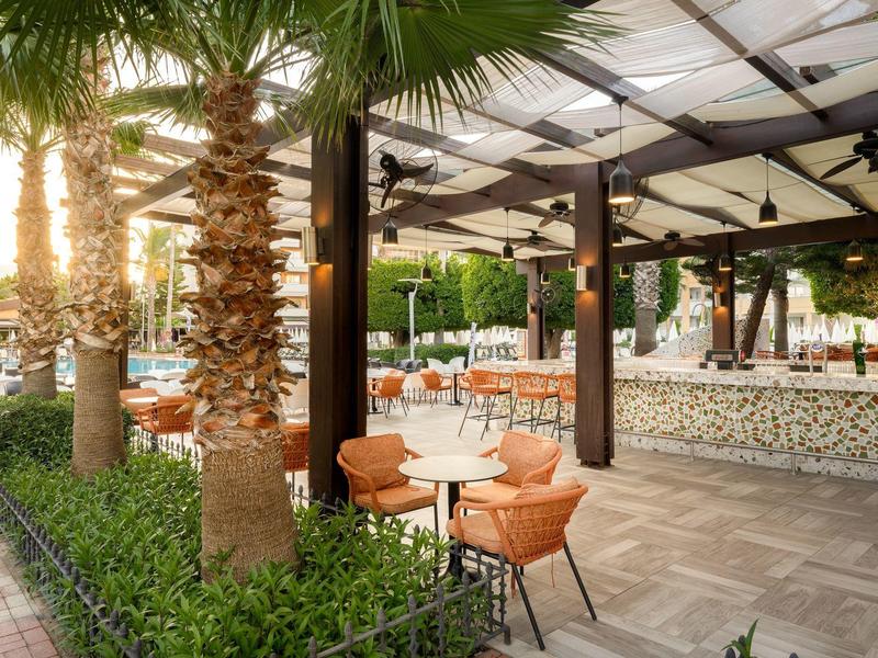 Außenbereich eines Cafés mit Palmen, orangen Stühlen und Tischen unter einem Glasdach.