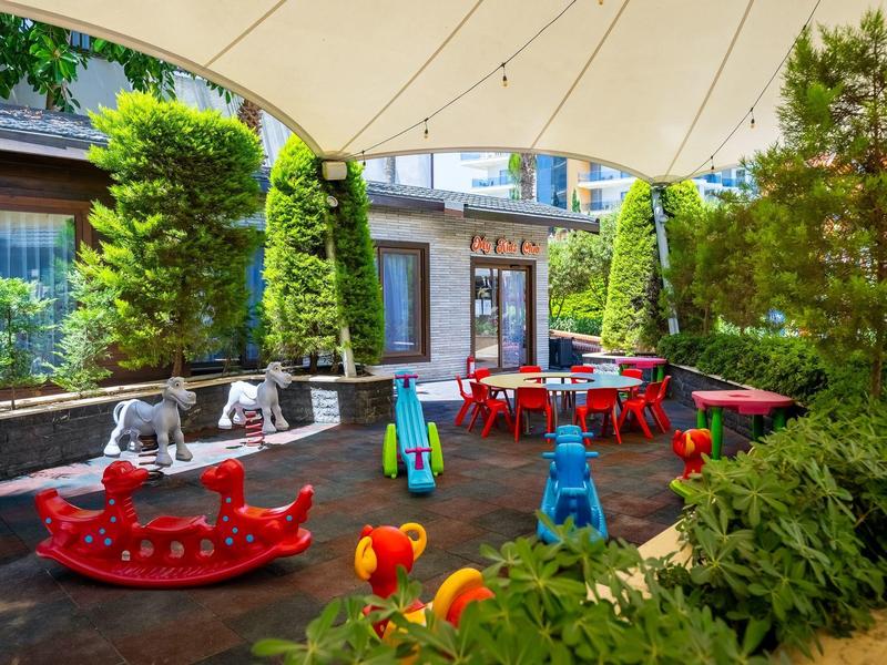 Bunter Kinderspielplatz mit Rutsche, Wippfiguren und Tischen unter Sonnensegel in begrüntem Innenhof.