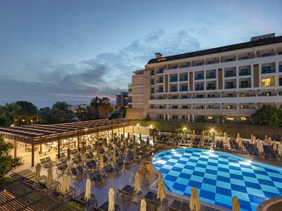 Toma nocturna de una piscina iluminada del hotel y área de terraza con mesas y sillas.