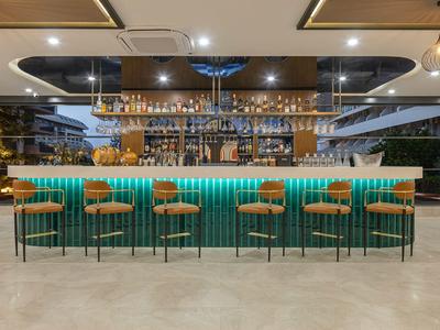Moderní hotelový bar s zeleným barem, dřevěnými stoličkami a skleněnými regály v pozadí.