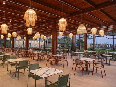 Venkovní restaurace s dřevěným stropem, útulným osvětlením a výhledem na tropické rostliny a moře.