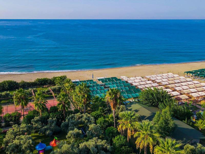 Complejo hotelero verde en la playa con sombrillas y mar azul al fondo.