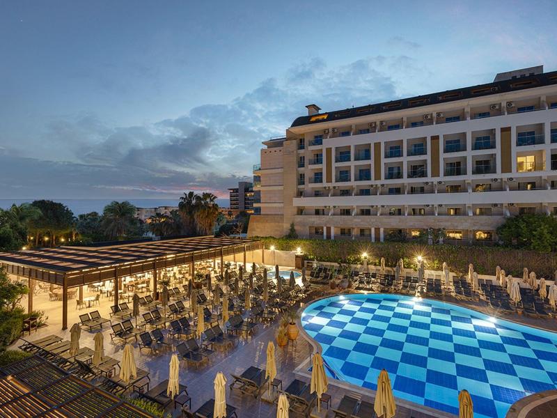 Toma nocturna de una piscina iluminada del hotel y área de terraza con mesas y sillas.
