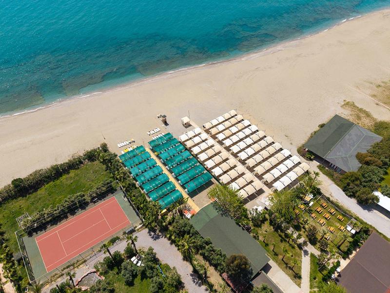 Vista aérea de una playa con filas de sombrillas blancas y azules, cancha de tenis y vegetación adyacente.