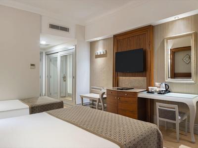 Moderně zařízený hotelový pokoj se dvěma postelemi, psacím stolem, televizí a stojací lampou.