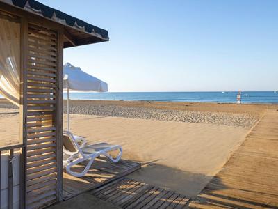 Pláž s lehátky, dřevěná cesta a moře v pozadí pod jasnou oblohou.