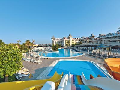 Venkovní hotelový bazén s tobogány, lehátky, stromy a jasnou modrou oblohou.