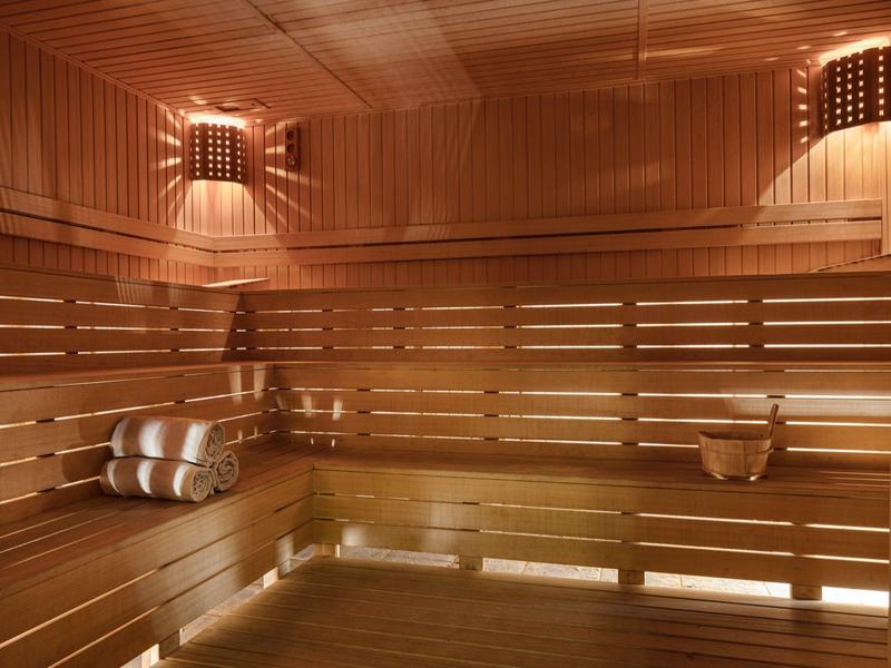 Světelná dřevěná sauna s lavicemi, ručníky a saunovým vědrem při teplém osvětlení.