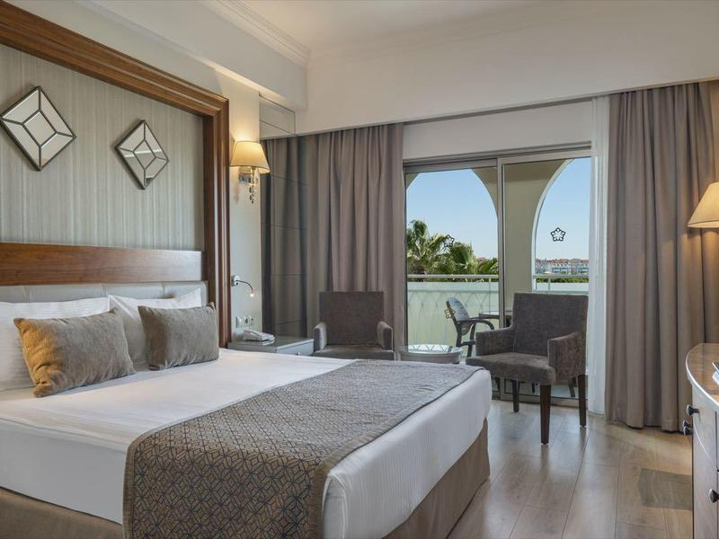 Světý hotelový pokoj s manželskou postelí, nočními stolky, posezením a výhledem na terasu se zahradou.