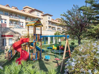 Parco giochi per bambini con scivolo e altalene davanti a un edificio alberghiero su più piani in una giornata di sole