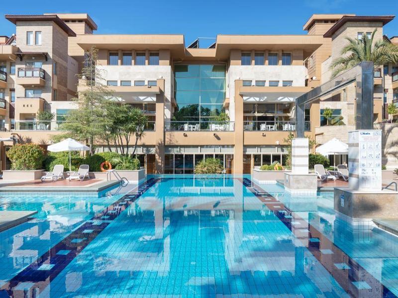 Grande piscina davanti a edificio alberghiero su più piani con ombrelloni e palme.