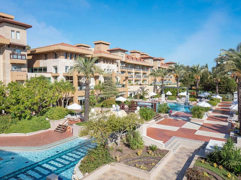 Hotel di lusso con piscina, palme e ombrelloni in una giornata soleggiata.
