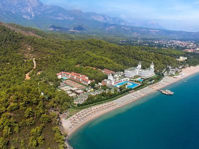 Luftaufnahme einer Küste mit Sandstrand, Hotels, Poolanlagen, bewaldeten Hügeln und Berglandschaft im Hintergrund.