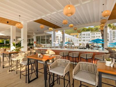 Modern hotel bar area with wooden and wicker furniture, open to the pool area with umbrellas.