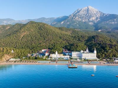 Beach hotel with white sand, blue sea, and mountains in the background under clear sky.