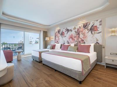 Bright hotel room with large bed, floral wall decor, and balcony overlooking the sea.