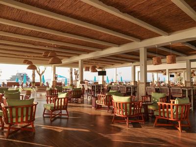 Geräumiger, offener Hotelrestaurantbereich mit Holzmöbeln und Blick aufs Meer bei Sonnenuntergang.