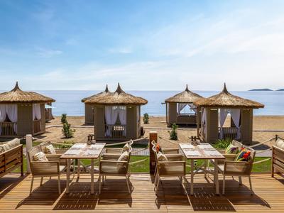 Strandhotel mit Holzterrasse, Tischen, Stühlen und gepolsterten Strandpavillons vor ruhigem Meer.