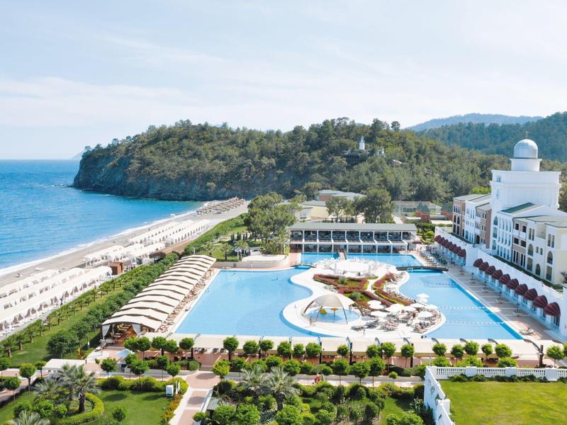 Großer Pool vor weißem Hotel, umgeben von grünen Hügeln, Sandstrand und blauem Meer im Hintergrund.