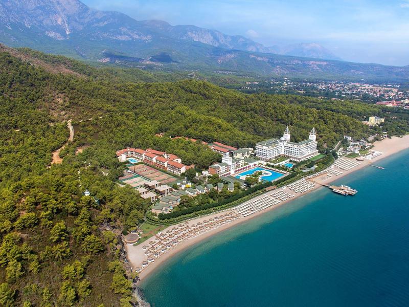 Luftaufnahme einer Küste mit Sandstrand, Hotels, Poolanlagen, bewaldeten Hügeln und Berglandschaft im Hintergrund.