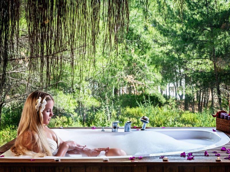 Frau entspannt in freistehender Badewanne im Freien, umgeben von grünen Bäumen und Blumen.