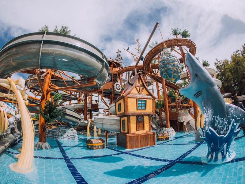 Bunter Wasserpark mit Rutschen, einem Hai-Modell und einer kleinen Holzhütte im blauen Pool.