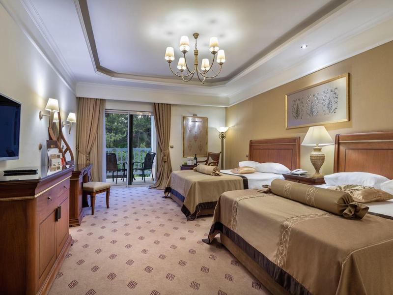 Hell eingerichtetes Hotelzimmer mit zwei Betten, Schreibtisch, Fernseher und Balkon mit Blick ins Grüne.