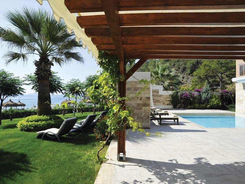 Haus mit Holzpergola, Pool und Liegestühlen, umgeben von grünem Rasen und Palmen am Meer.