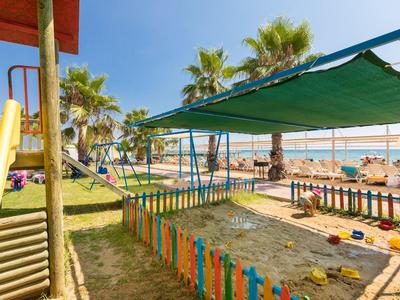 Parco giochi per bambini con sabbionaia e scivolo sulla spiaggia, coperto e con palme sullo sfondo.