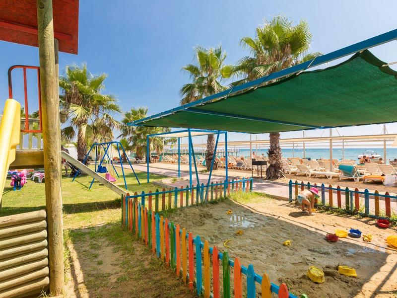 Parco giochi per bambini con sabbionaia e scivolo sulla spiaggia, coperto e con palme sullo sfondo.