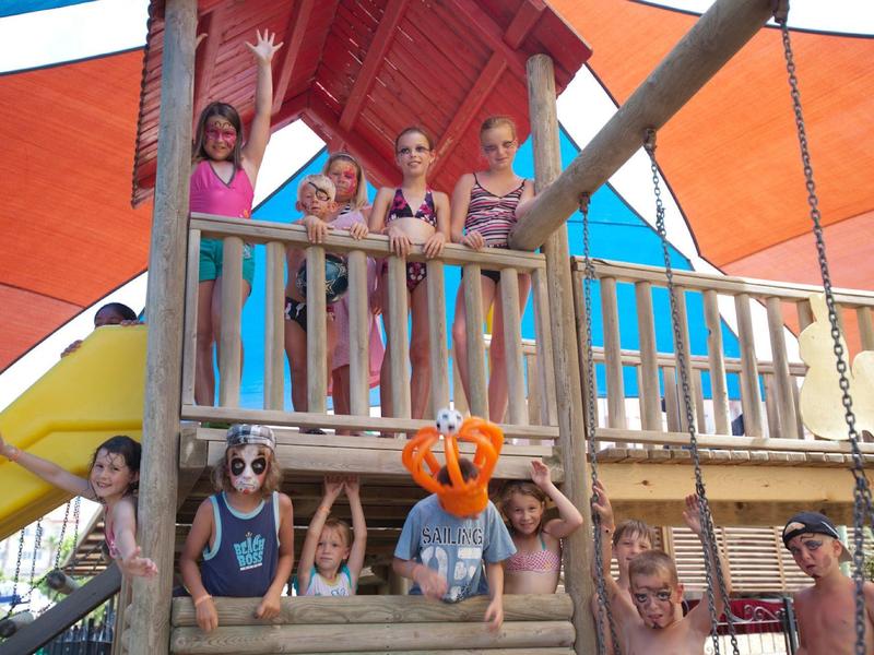 Bambini che giocano su un parco giochi in legno con scivoli sotto tende arancioni.