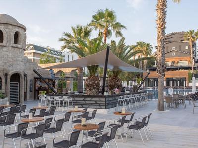Outdoor-Café mit Stühlen, Tischen, Palmengarten und mediterraner Architektur bei klarem Himmel.