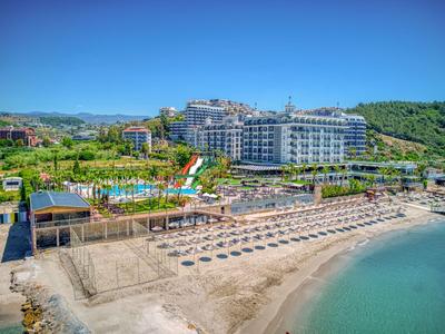 Hotelanlage an einem Sandstrand mit Liegen, Pool und Hügeln im Hintergrund.