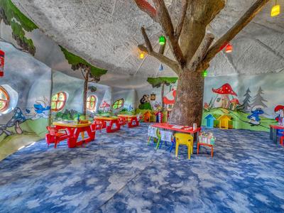 Kinderspielzimmer mit bunten Tischen und Bäumen, Wände mit Spiel- und Landschaftsmalereien dekoriert.