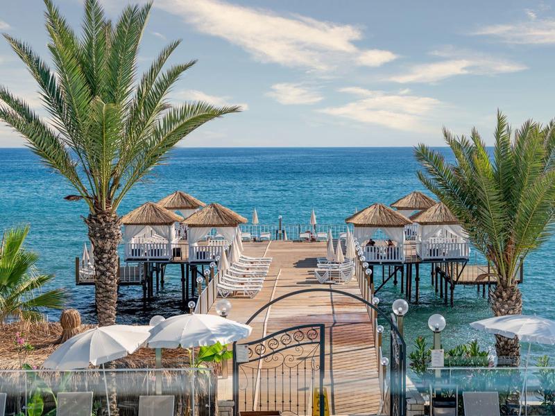 Jetée en bois bordée de palmiers menant à un pavillon sur la mer bleue sous un ciel nuageux.