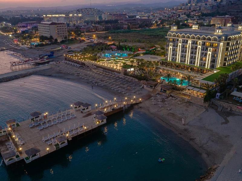 Vue aérienne d'une station balnéaire avec piscine, jetée et ville en arrière-plan au coucher du soleil.