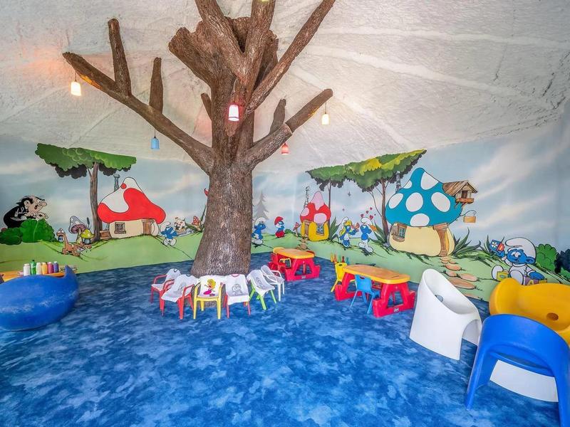 Espace de jeu coloré pour enfants avec fresque d'arbre et tables colorées sur sol bleu.