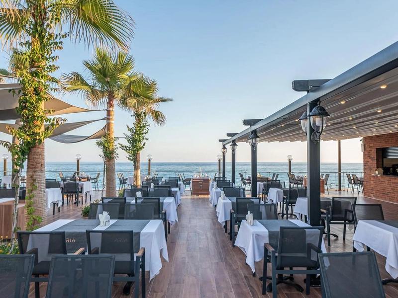 Restaurant en plein air avec nappes blanches, vue sur la mer et palmiers sous un ciel dégagé.