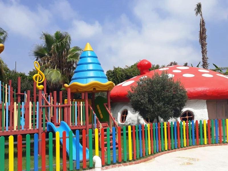 Aire de jeux colorée avec maison champignon, toboggan et clôture vive sous ciel bleu.