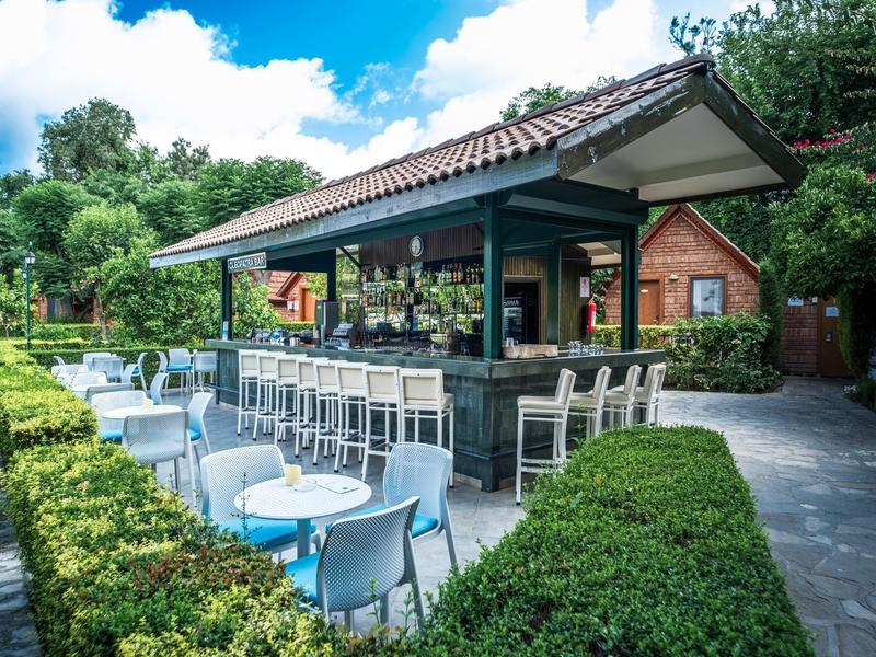 Buitenkant bar met witte stoelen en tafels omgeven door groene hagen en bomen.
