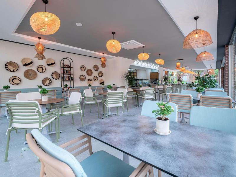 Modern café met planten, hanglampen en lichte stoelen bij grote ramen.