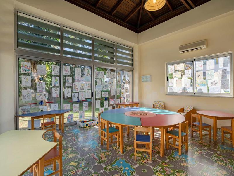 Stanza luminosa con tavoli rotondi di legno, sedie, grandi finestre e poster alle pareti.