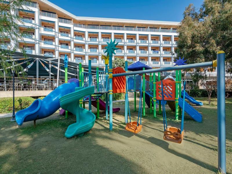 Parco giochi colorato con altalene e scivoli davanti a un hotel moderno.